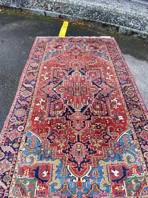 Un tapis persan orné d'un motif géométrique complexe aux tons rouge, bleu et jaune repose sur une surface en béton humide. Ce motif complexe comprend des motifs floraux et abstraits symétriques : une véritable élégance. Une ligne jaune et du gravier sont visibles en arrière-plan.
