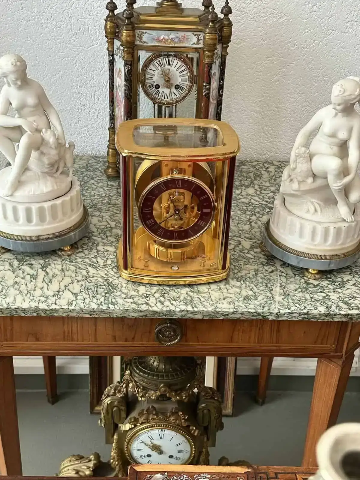 Sur une table en marbre de Bienne, une pièce décorative présente une horloge dorée au boîtier transparent et deux sculptures blanches représentant des personnages assis. À l'arrière-plan, une horloge vintage ornée de détails ornementaux. Une horloge en bois est partiellement visible en dessous.