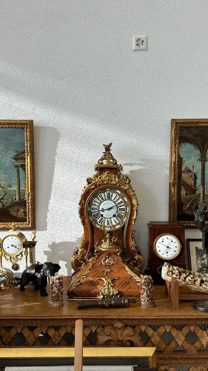 Une table en bois présente des horloges anciennes, des figurines, des assiettes décoratives et des objets sculptés de Nyon. Au-dessus, deux tableaux de paysages encadrés sont accrochés à un mur blanc, éclairés par la lumière du soleil.