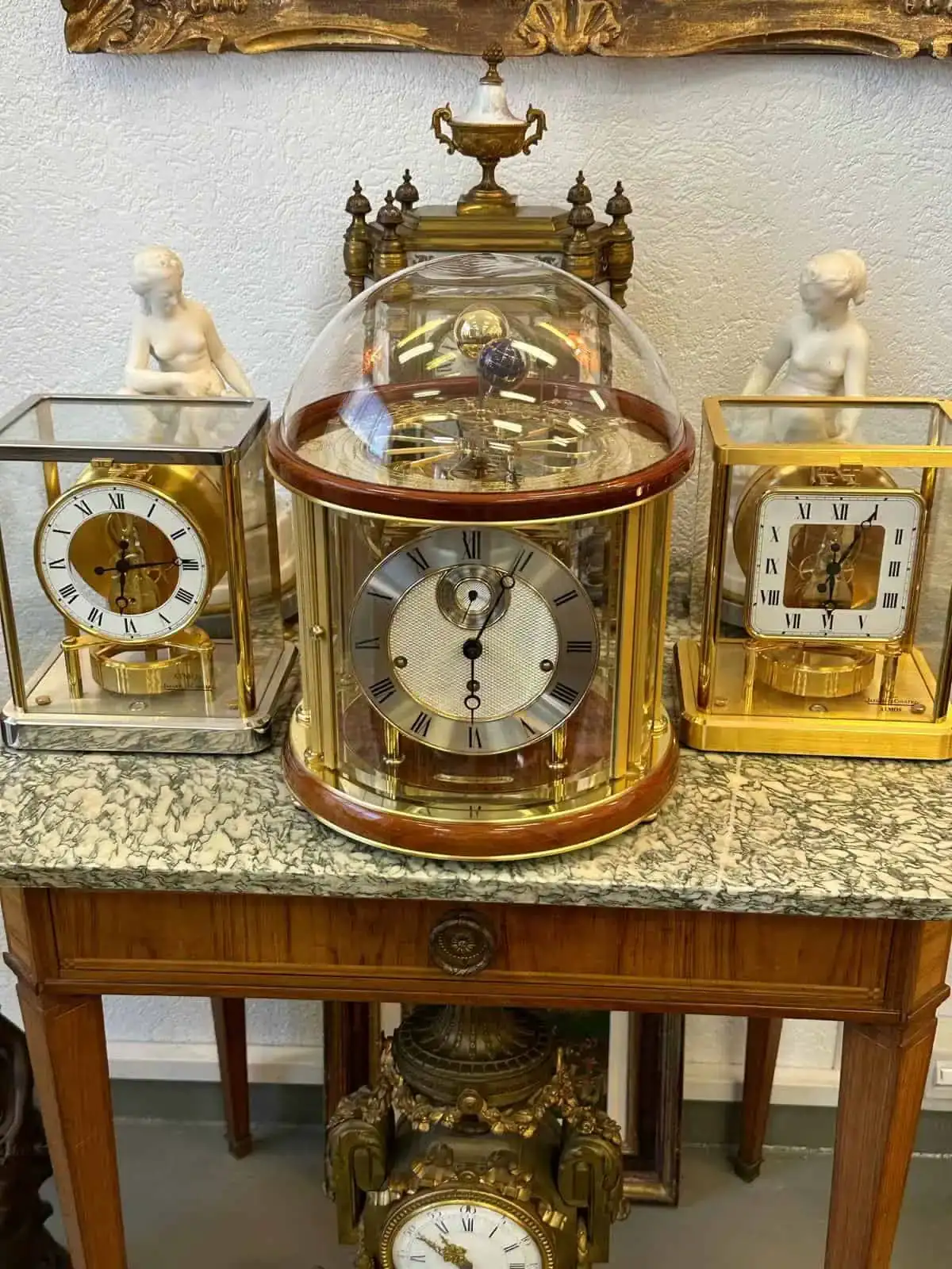 À Bienne, une table en bois au plateau de marbre présente trois horloges ouvragées. L'horloge centrale, encastrée dans une coupole de verre, dévoile ses mécanismes complexes, tandis que deux horloges à chiffres romains, encadrées d'or, se dressent de chaque côté. Statues et objets décoratifs rehaussent l'élégant décor.