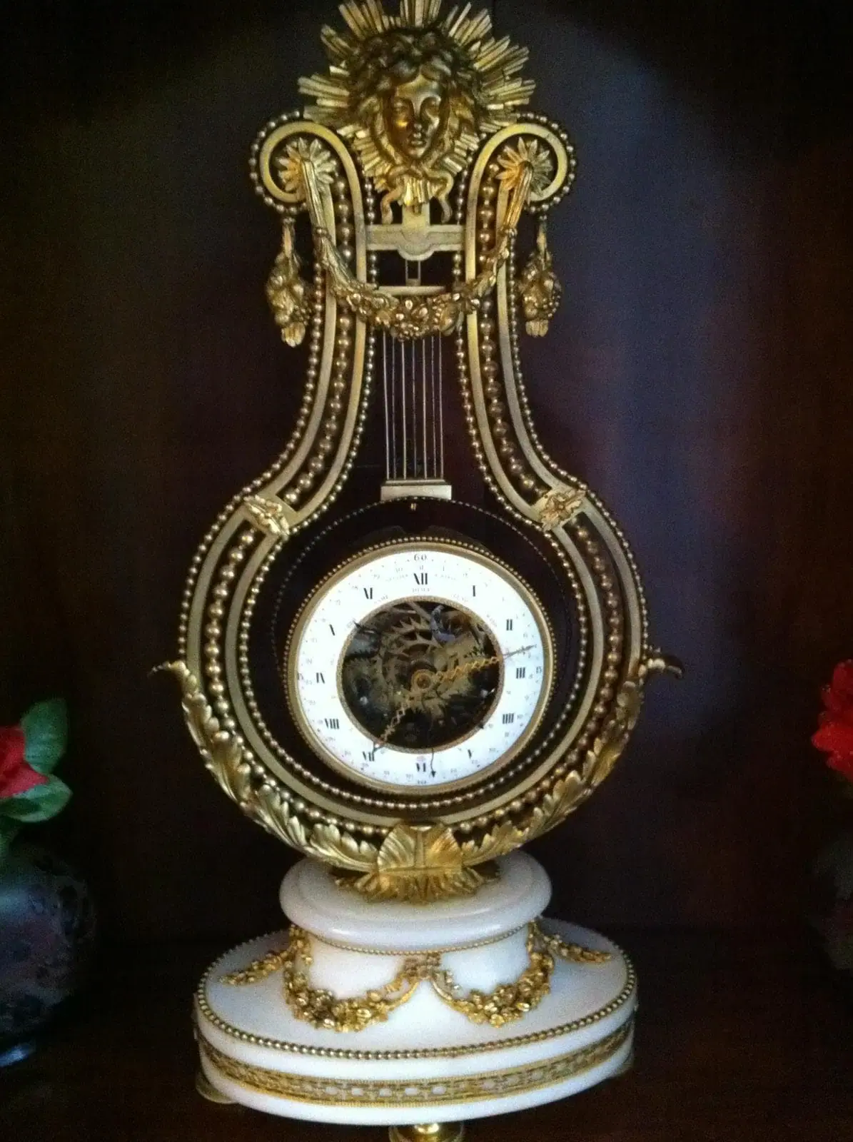 Cette horloge ornée en forme de lyre, inspirée de l'élégance lausannoise, présente de délicats détails dorés et une base blanche ornée de guirlandes décoratives. Des chiffres romains ornent le cadran, couronné d'un soleil et d'un cadran dorés. Des fleurs rouges accentuent délicatement ses côtés.