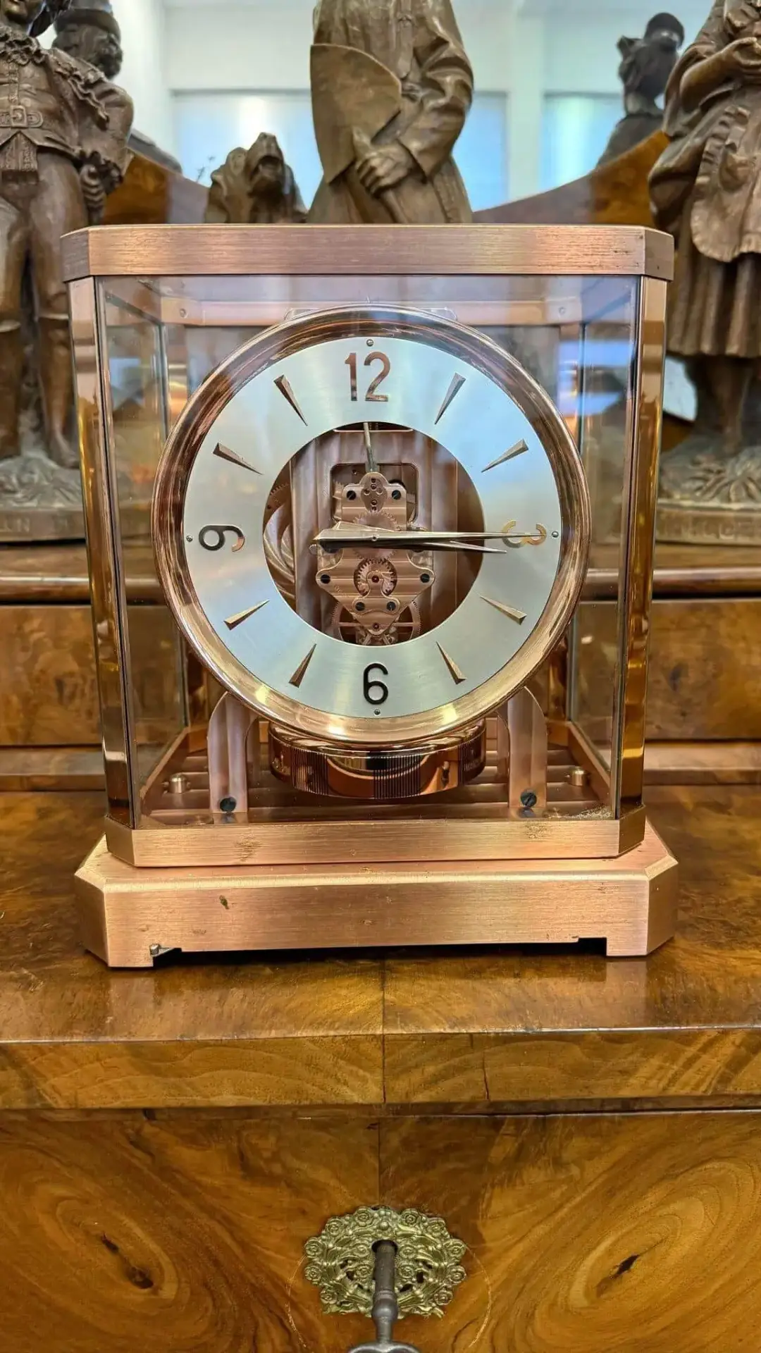 Une horloge de cheminée vintage Sion en verre et cuivre repose sur une surface en bois poli, son mécanisme complexe étant visible à l'intérieur. Des figurines en bronze sont exposées en arrière-plan.