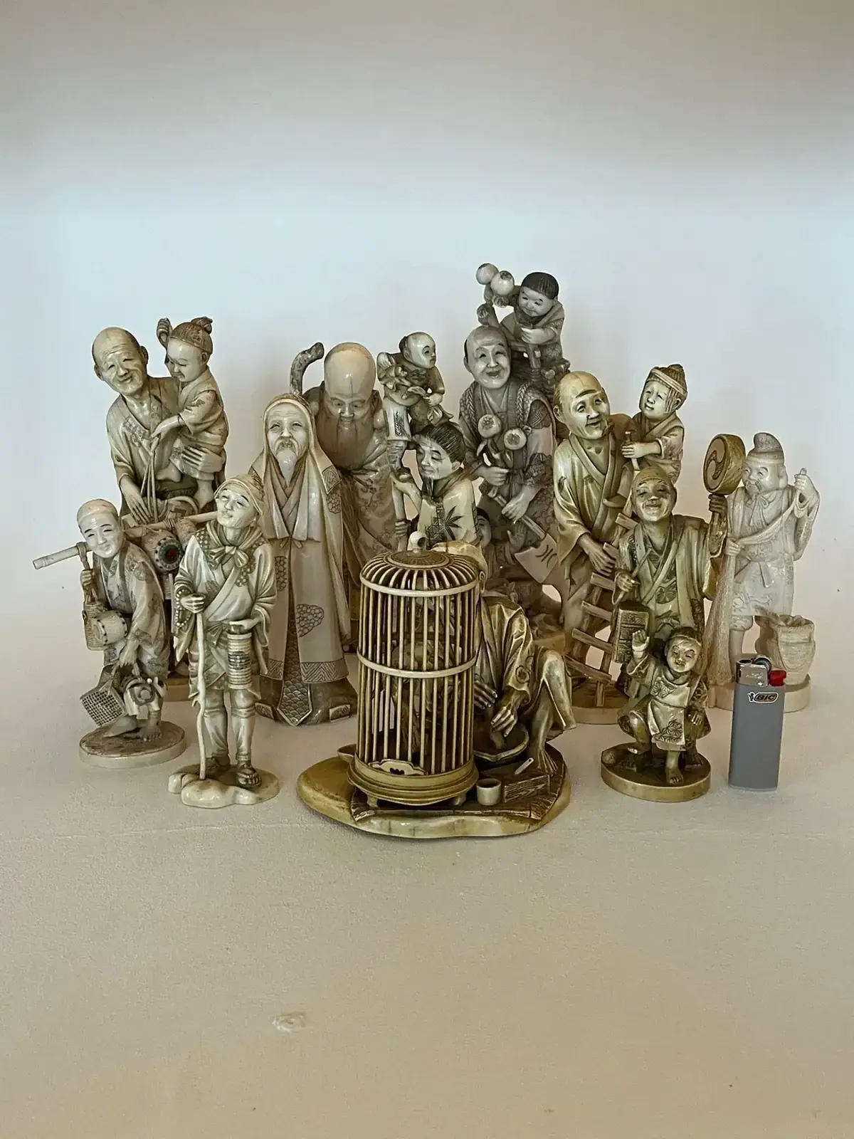 Un groupe de figurines en ivoire ou en os de Sion finement sculptées, représentant diverses personnes en vêtements traditionnels, sont rassemblées autour d'une cage à oiseaux centrale, avec un briquet à droite pour l'échelle.