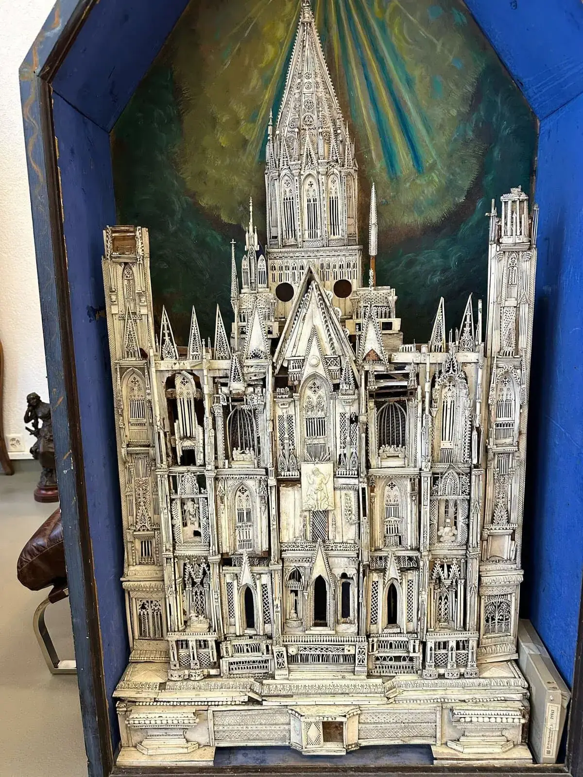 Maquette miniature détaillée d'une cathédrale de style gothique, finement sculptée en ivoire ou matériau similaire, présentée dans une vitrine en bois bleu. Le fond peint en vert et jaune évoque des rayons de lumière, rappelant le talent artistique de Montreux.