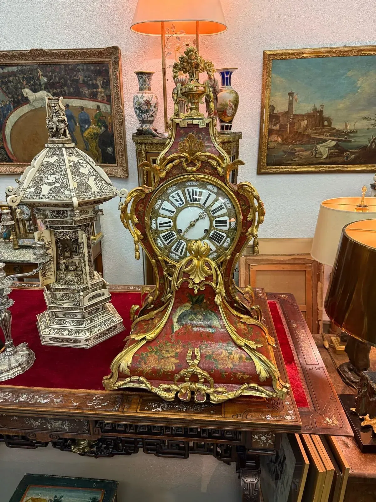 Une horloge antique Nyon ornée avec des accents dorés et des chiffres romains se trouve sur une table en velours rouge, entourée de lampes vintage, de vases et de peintures encadrées dans un magasin d'antiquités.