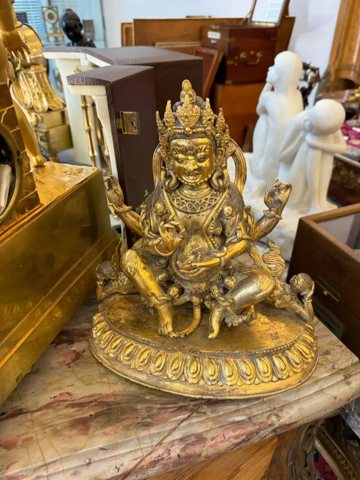 Une statue dorée détaillée d'une divinité à plusieurs bras est assise en tailleur sur un piédestal orné dans un magasin d'antiquités de Berne, entourée de divers objets et boîtes sur une surface en marbre.