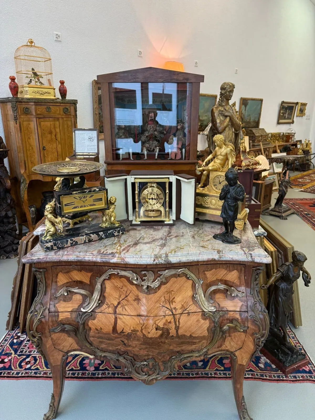 Une vitrine de magasin d'antiquités présente un coffre en bois orné avec une scène pastorale peinte de Lucerne, des statues en bronze, des horloges, une cage à oiseaux dorée et des œuvres d'art encadrées, le tout magnifiquement disposé dans une pièce bien éclairée.