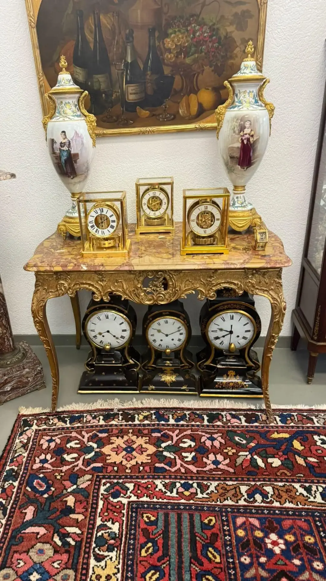 Une table ancienne décorative de Lucerne présente des horloges ouvragées et deux grands vases en porcelaine. D'autres horloges reposent sur une étagère inférieure, entourées d'un tapis aux motifs complexes, de tableaux classiques et d'un mobilier élégant.