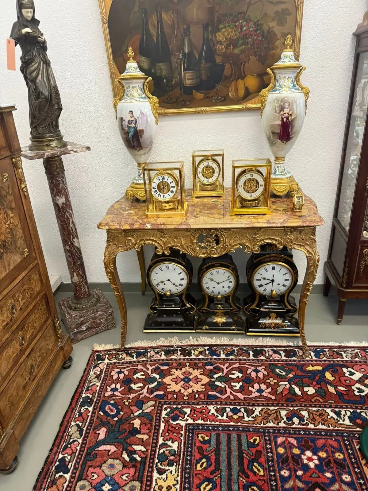 Une table neuchâteloise ancienne et ornée, ornée d'horloges décoratives et de grands vases en porcelaine, trône contre un mur. Un tapis à motifs colorés recouvre le sol et une nature morte est suspendue au-dessus de la table.