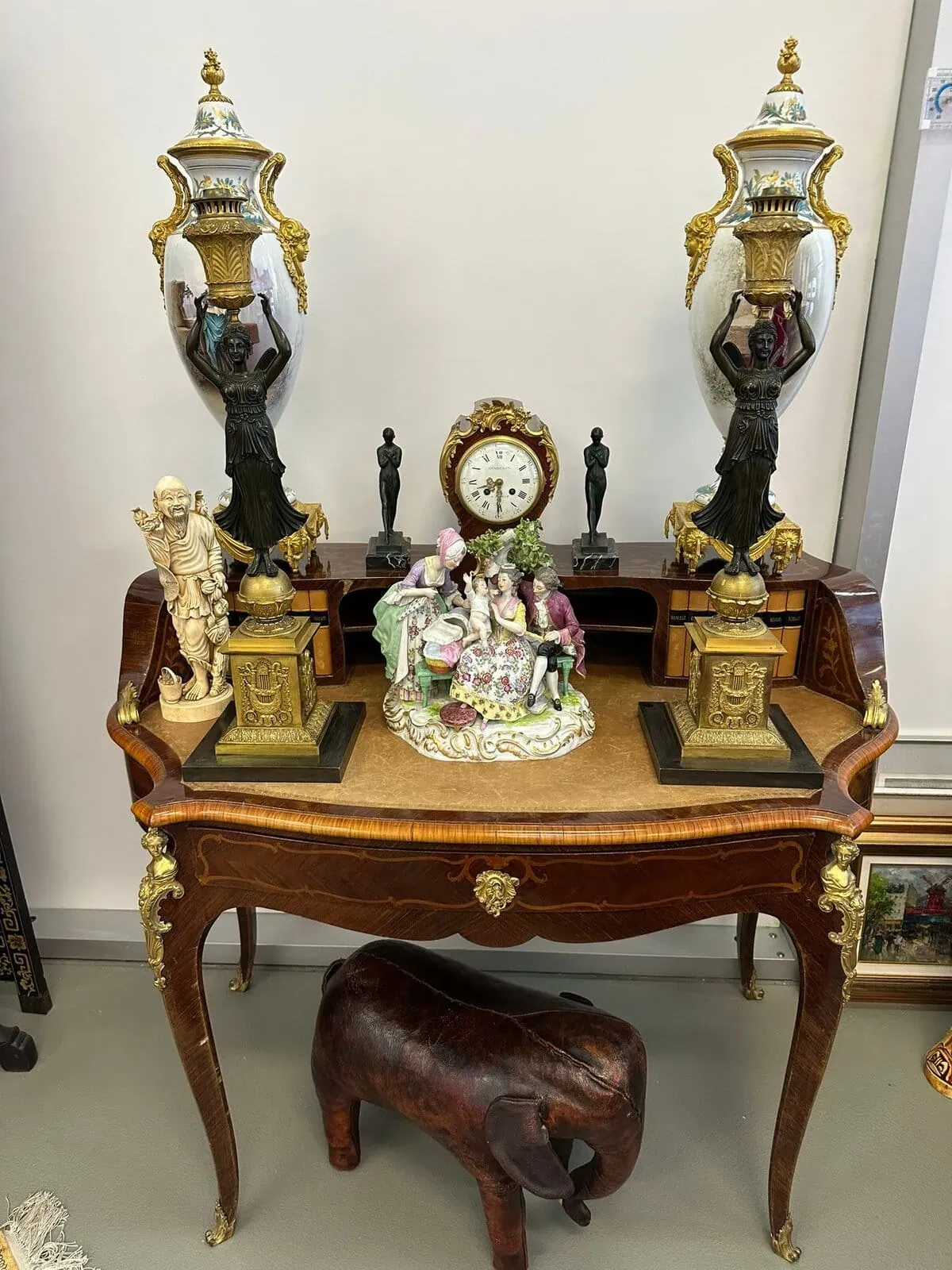 Une table en bois ornée, rappelant l'artisanat de La Chaux-de-Fonds, présente deux grandes urnes décoratives, un groupe de figurines en porcelaine, une petite horloge, des statues et une figure en ivoire sculpté. Un éléphant en bois trône au sol, sous la table.