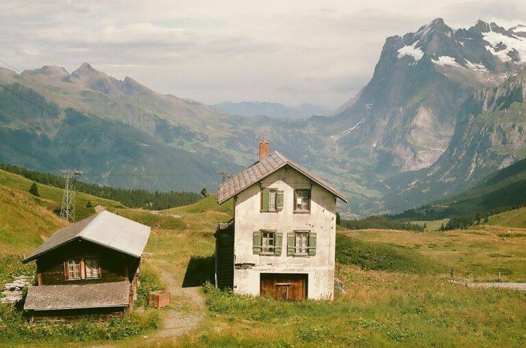 Deux maisons rustiques se dressent sur une colline verdoyante avec une chaîne de montagnes spectaculaire et des sommets enneigés en arrière-plan sous un ciel partiellement nuageux.