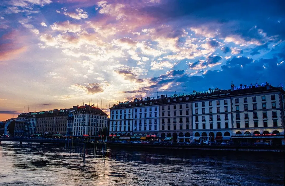 Une rangée de grands bâtiments aux enseignes lumineuses borde une rivière sous un coucher de soleil vibrant et coloré, parsemé de nuages spectaculaires. Les reflets des bâtiments et des lumières scintillent sur l'eau.