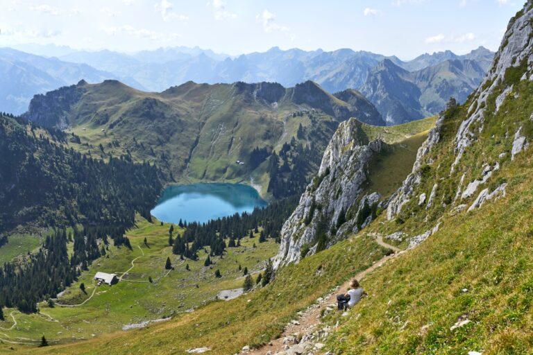 Un randonneur est assis sur une pente de montagne herbeuse surplombant un lac alpin turquoise entouré de collines boisées et de sommets escarpés lointains sous un ciel partiellement nuageux.