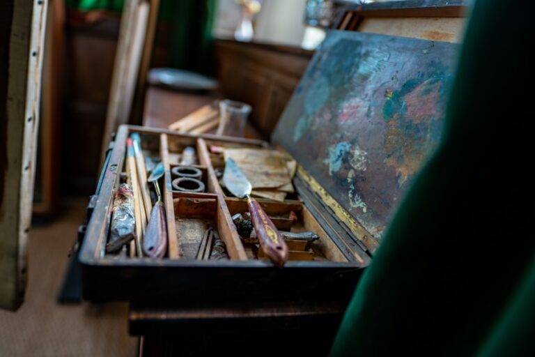 Une boîte de peinture en bois ouverte présente des pinceaux, des couteaux à palette et du matériel de peinture usagés dans plusieurs compartiments, avec une palette maculée de peinture posée au-dessus des outils. L'arrière-plan est légèrement flou.