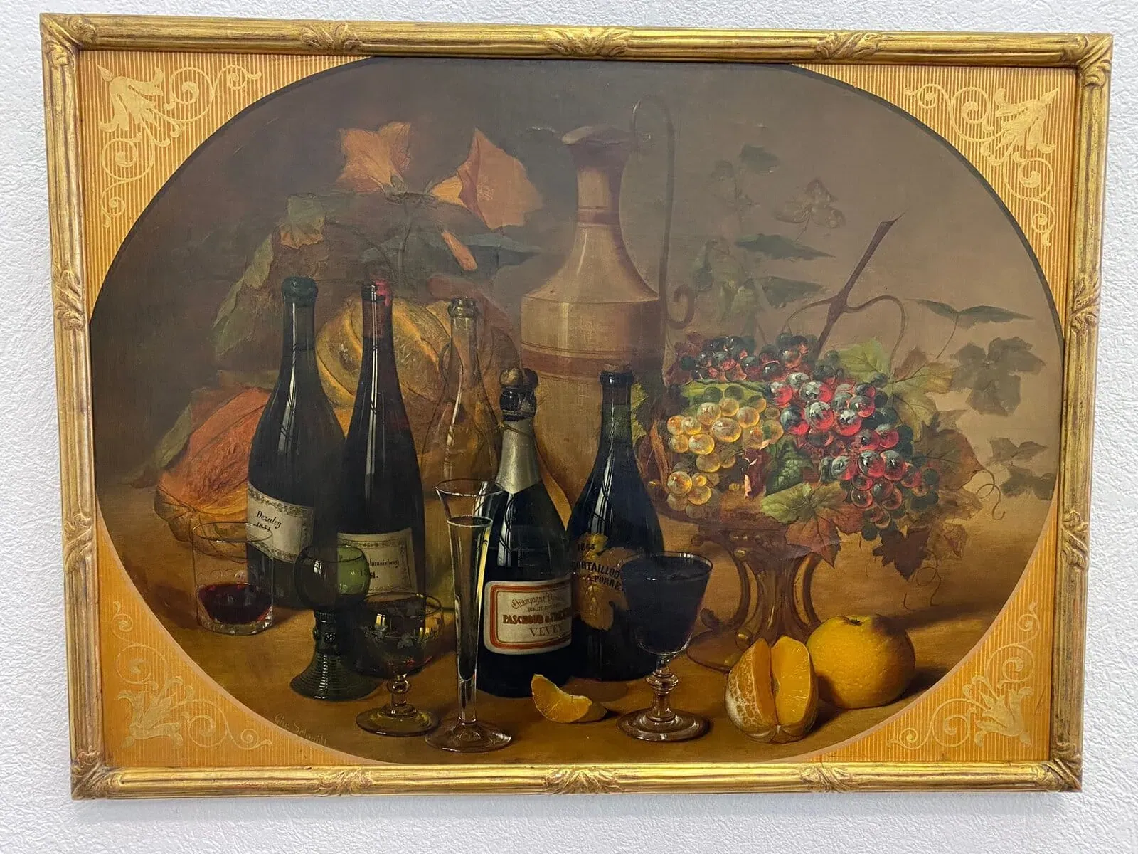 Une nature morte représentant des bouteilles de vin, des verres, une carafe, des fruits dont des oranges et des raisins, et une citrouille, le tout disposé sur une table avec un fond décoratif et un cadre doré orné.