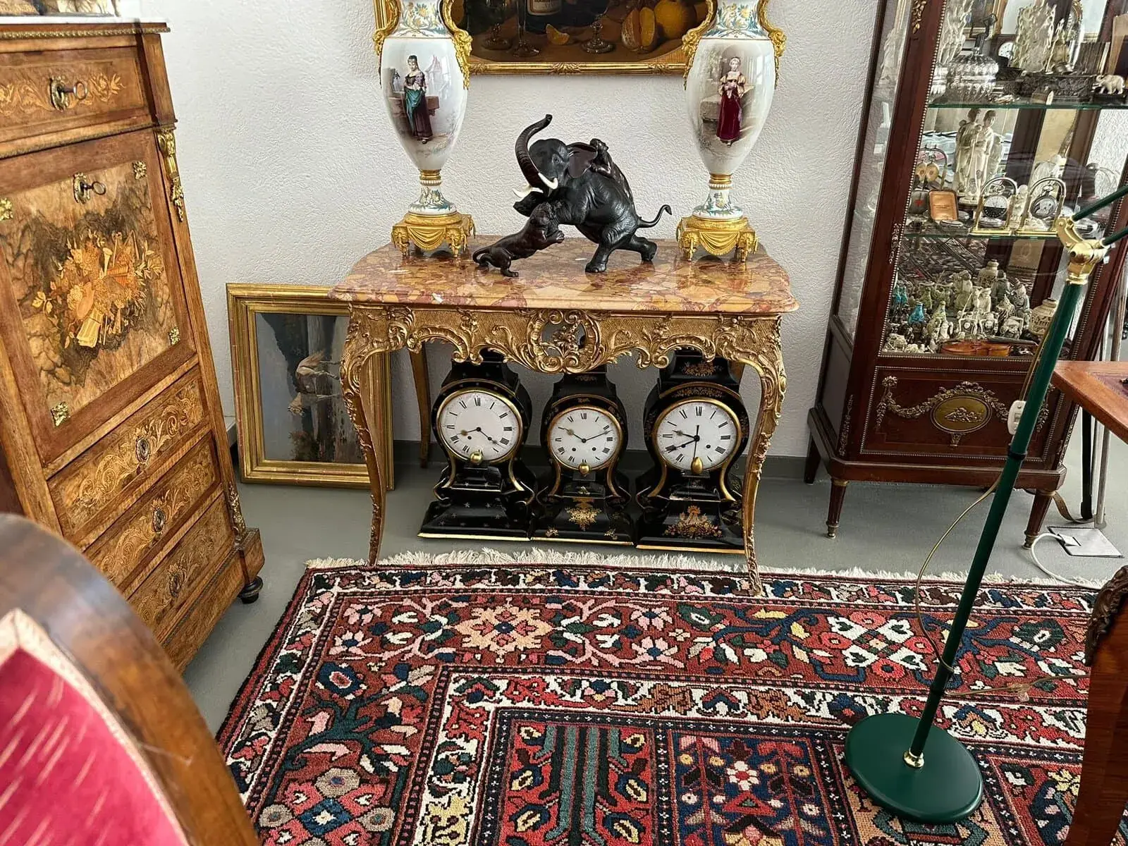 Une table ornée avec trois horloges anciennes en dessous, surmontée de deux vases décoratifs et d'une figurine d'éléphant noir, se trouve à proximité de meubles vintage et d'un tapis à motifs colorés dans une pièce pleine d'antiquités.