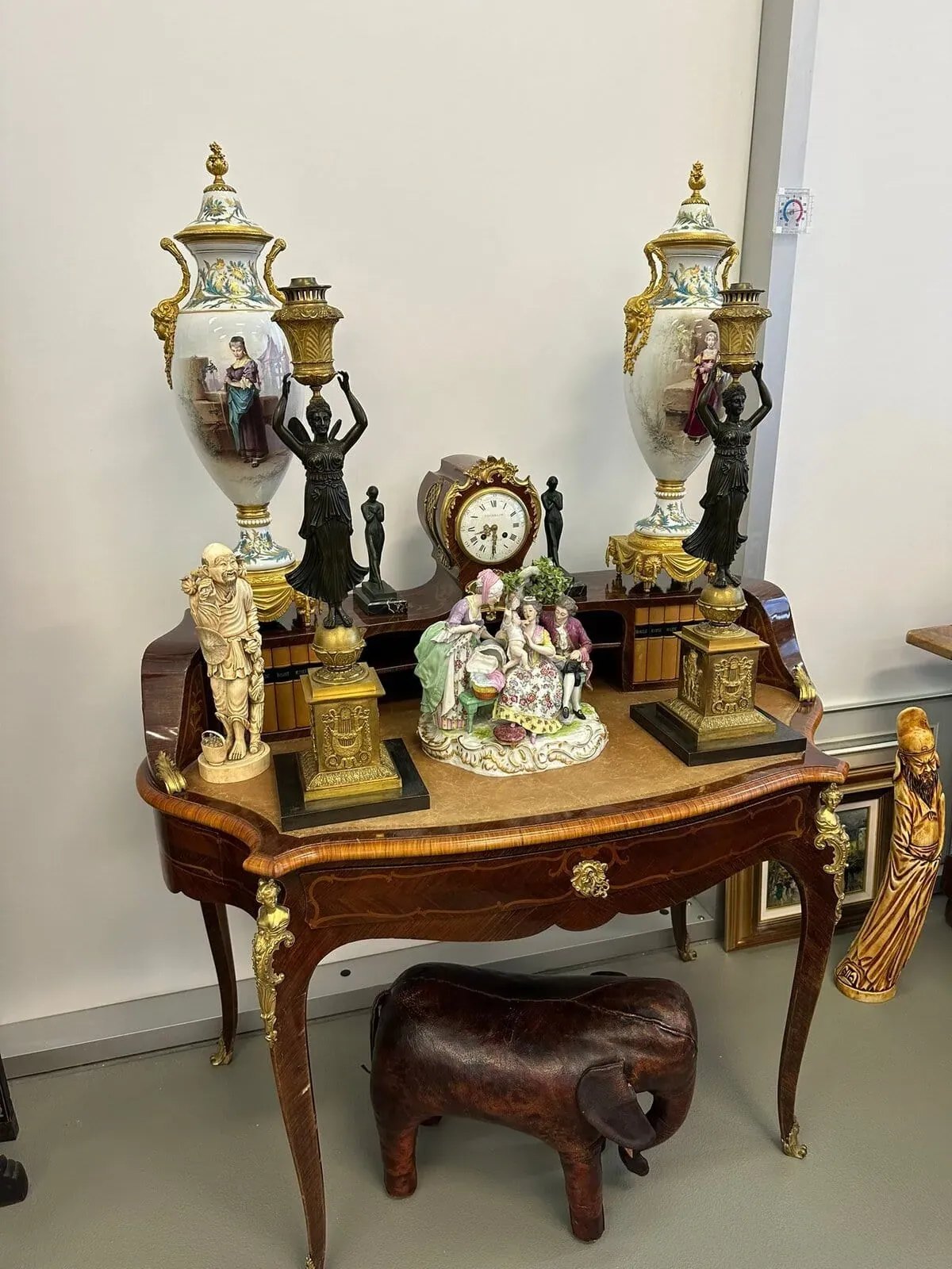 Une table en bois ornée présente des objets décoratifs, dont deux grandes urnes, une horloge, deux statues en bronze contenant des lampes, des figurines en porcelaine, un éléphant sculpté sous la table et des sculptures plus petites.