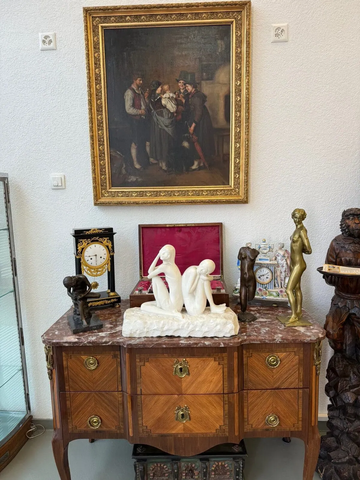 Une commode en bois présente des horloges anciennes, une boîte à bijoux rouge et plusieurs sculptures. Au-dessus est accroché un tableau encadré représentant des personnes en vêtements d'époque en train de converser. La scène est élégante et vintage.
