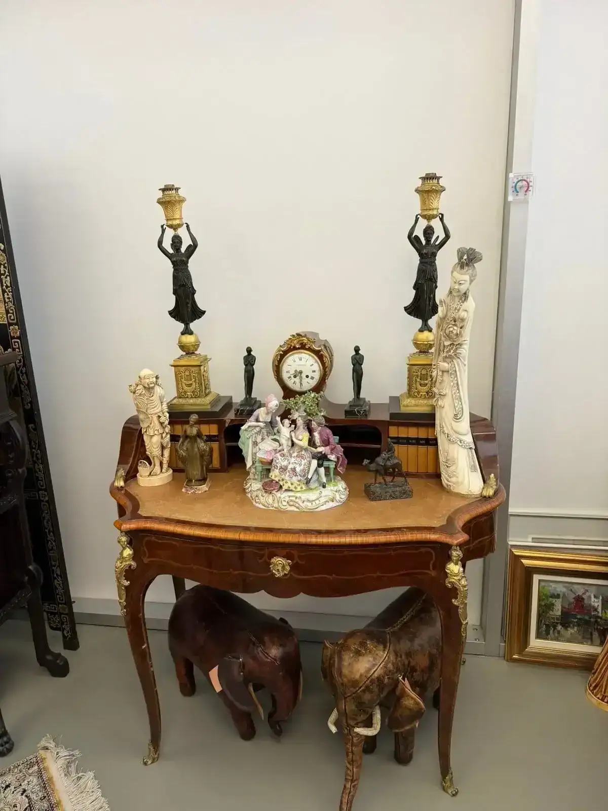 Une table en bois ornée présente divers objets décoratifs, dont deux chandeliers féminins en bronze, une horloge, des figurines en porcelaine, de petites sculptures d'éléphants et deux éléphants en bois sous la table. Des tableaux sont adossés au mur à proximité.
