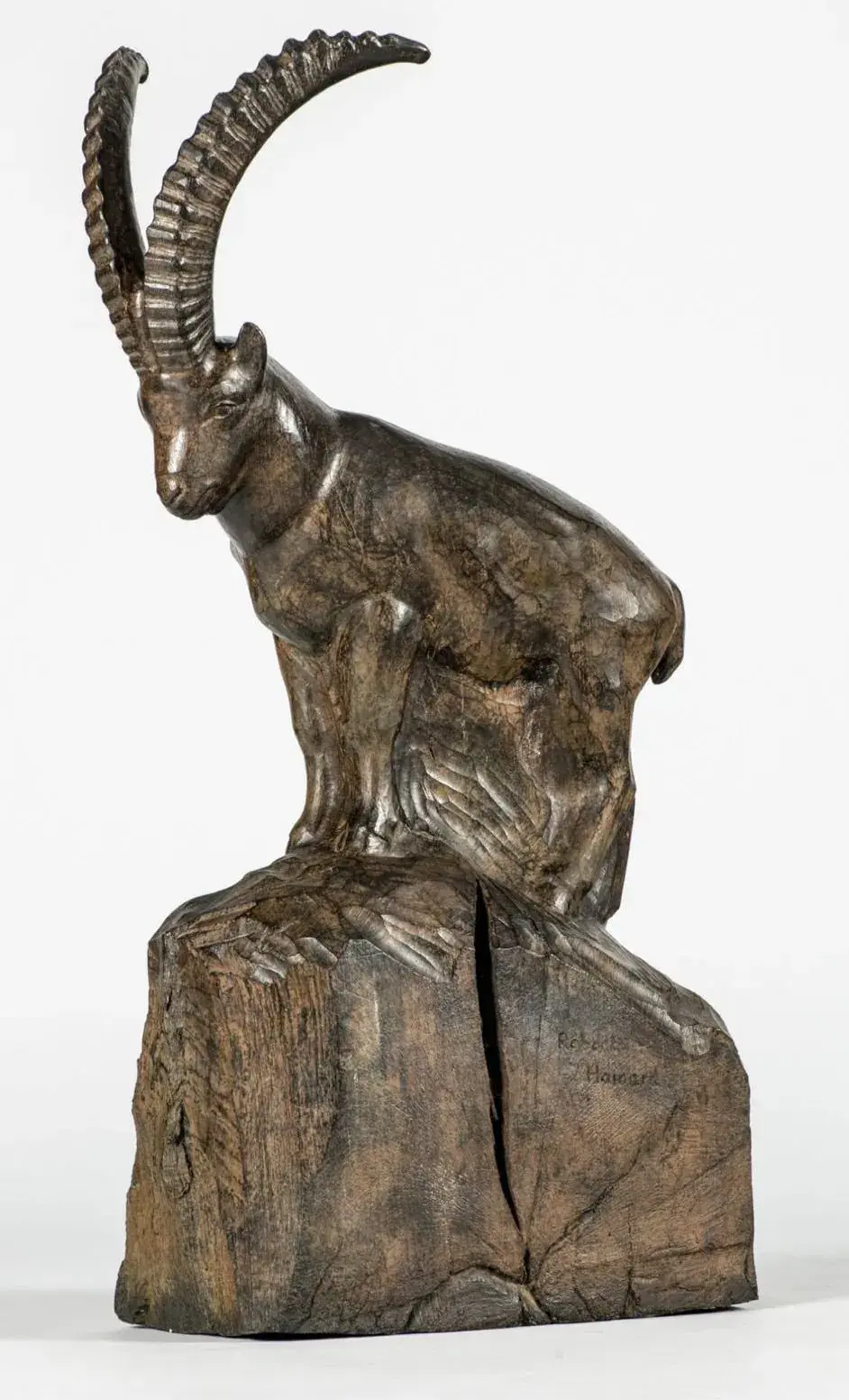 Une sculpture en bois d'un bouquetin aux cornes incurvées repose sur une base rocheuse texturée. Le bois est sombre et poli, avec des veines visibles et une fissure dans la base.