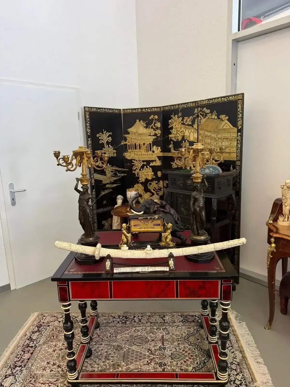 Une table décorative avec des candélabres ornés, une défense en ivoire incurvée et une petite statue est posée sur un tapis à motifs devant un paravent noir et or de style asiatique aux motifs complexes.