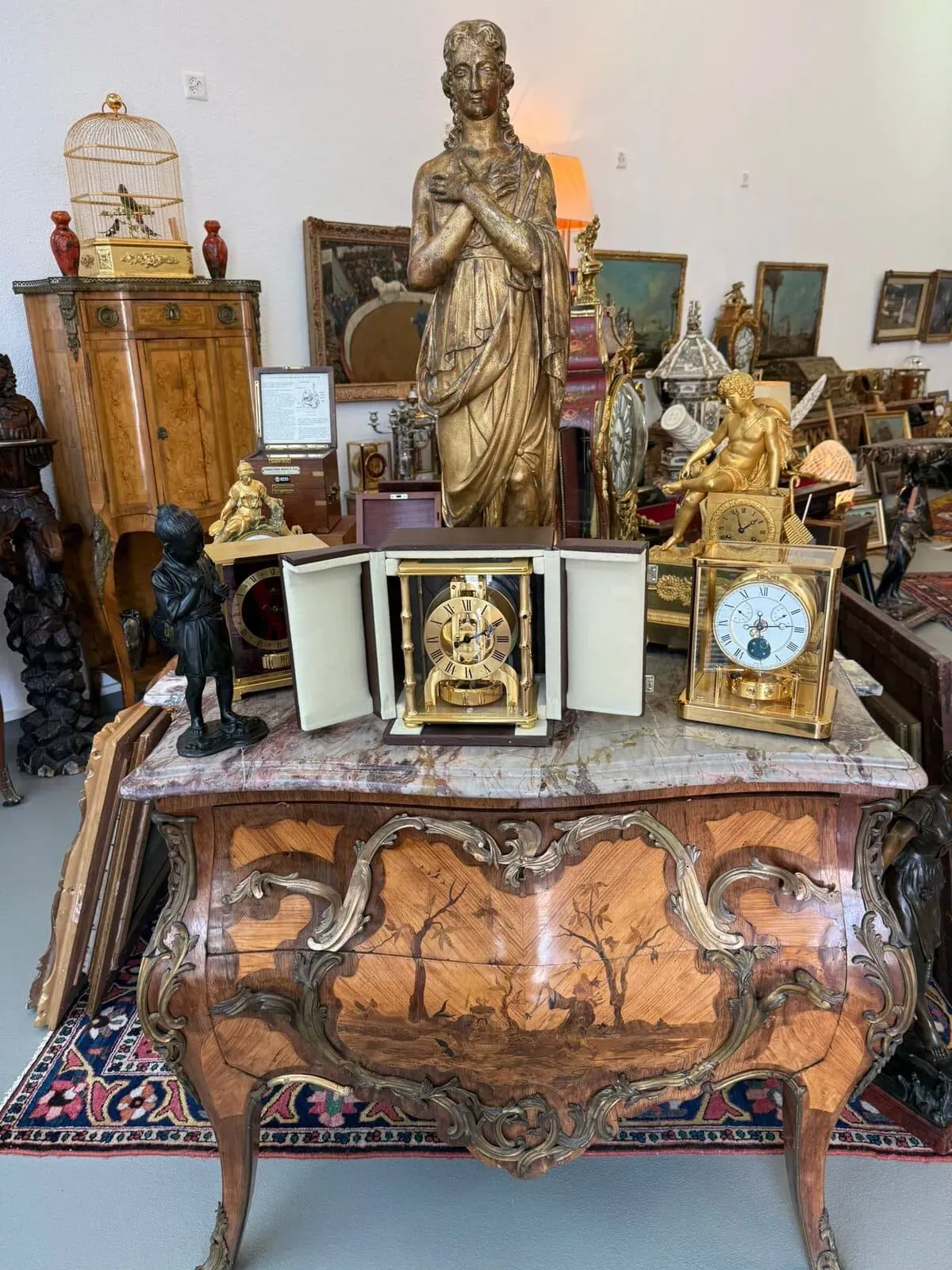 Un coffre en bois orné d'arbres sculptés contient des horloges anciennes, des statues en or et en bronze et des objets de décoration antiques. Une cage à oiseaux et des œuvres d'art encadrées sont visibles dans une pièce remplie d'objets de collection éclectiques et historiques.