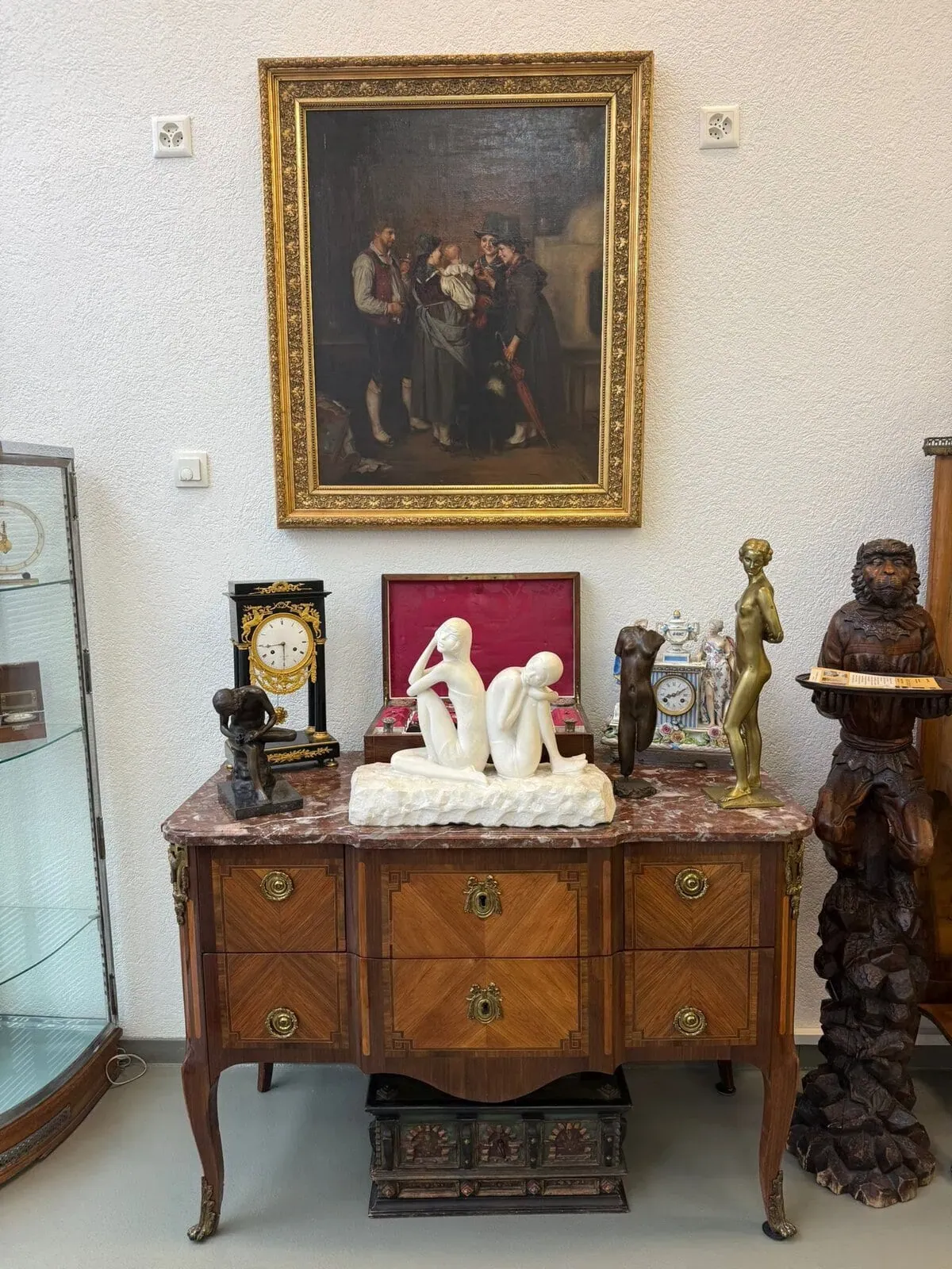 Une commode en bois avec un plateau en marbre contient des sculptures, des horloges et des objets décoratifs ; au-dessus, un tableau encadré représentant trois personnes est accroché à un mur blanc. Une vitrine en verre se trouve à gauche.