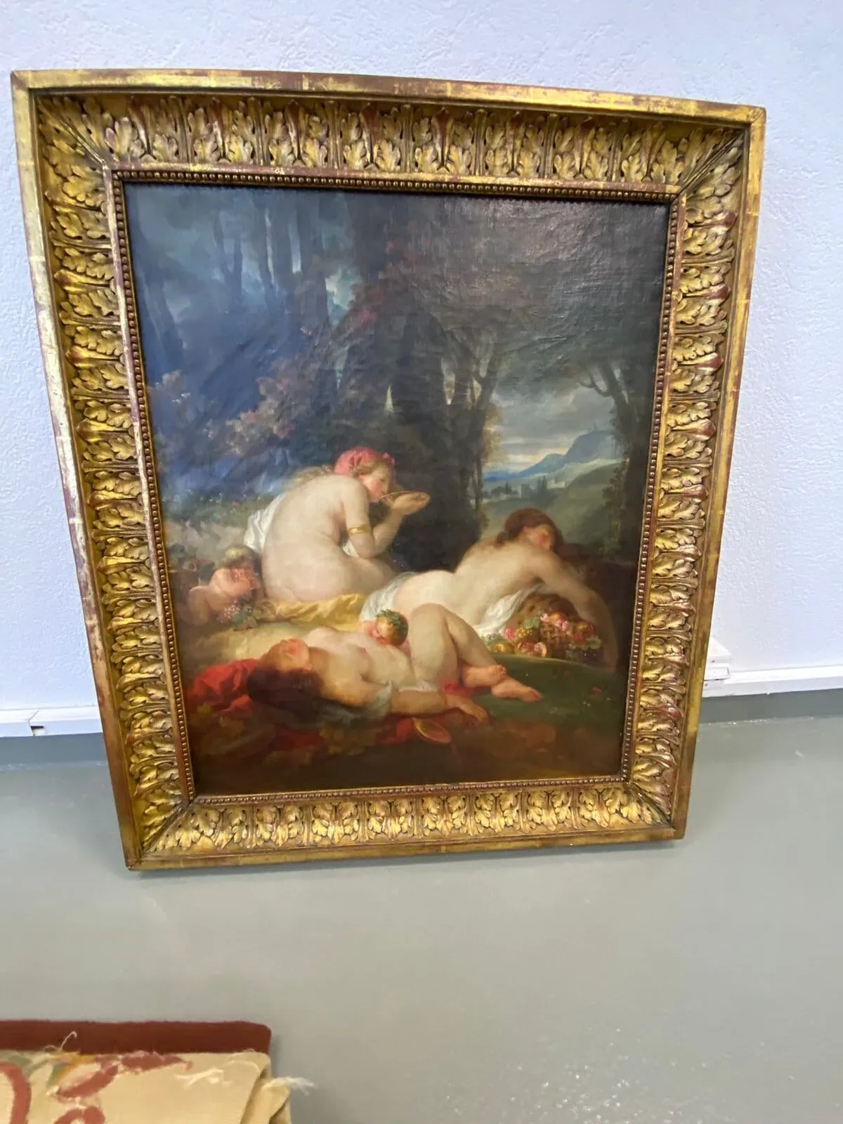 Tableau encadré représentant trois personnages nus allongés sur l'herbe dans un paysage forestier, avec une corbeille de fruits à proximité. Le cadre doré est adossé à un mur clair.