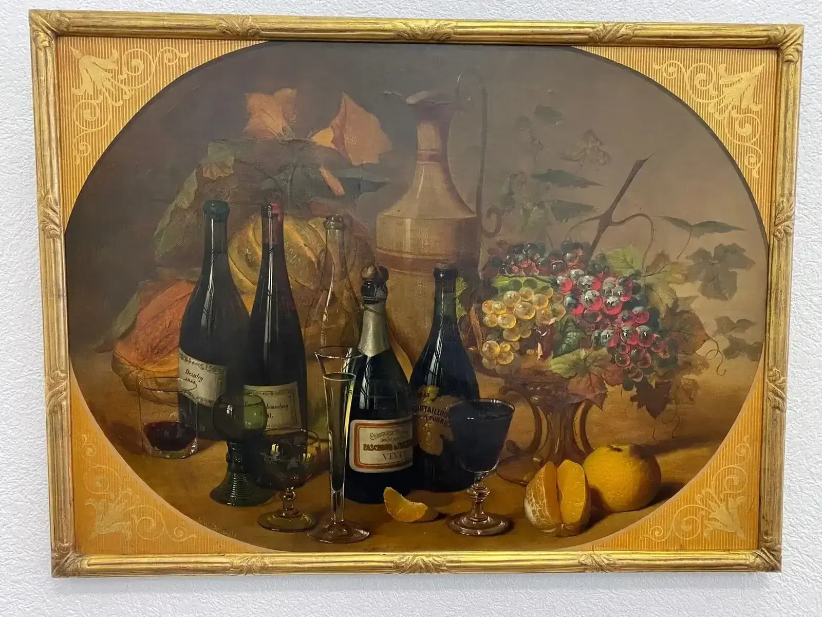 Une nature morte encadrée représentant des bouteilles de vin, des verres, une carafe, des fruits assortis, des raisins et des oranges, le tout disposé sur une table avec des feuilles en arrière-plan. Le cadre est orné et doré.