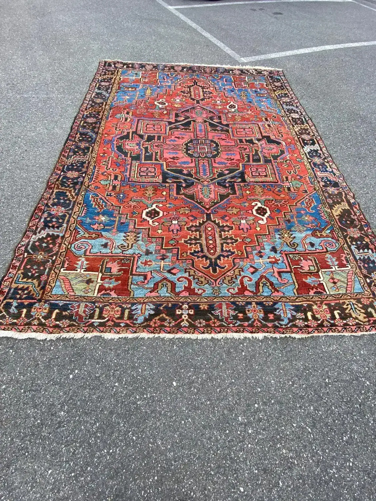 Un grand tapis oriental Zoug aux motifs géométriques et floraux complexes de couleur rouge, bleue, noire, ivoire et verte est posé à plat sur une surface asphaltée grise, avec des lignes de parking blanches visibles à l'arrière-plan.