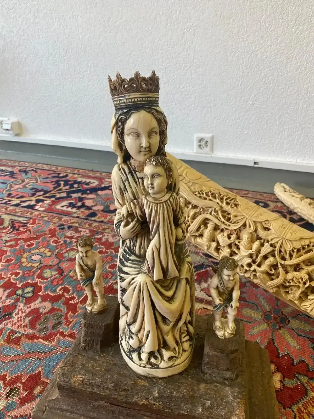 Une statue sculptée de couleur ivoire représentant une femme couronnée tenant un enfant, peut-être la Vierge à l'Enfant, repose sur une base en bois. Deux personnages agenouillés plus petits la flanquent. À l'arrière-plan, un tapis orné du canton de Zoug et des boiseries détaillées se trouvent près d'un mur blanc.