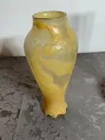 Un grand vase en verre jaune avec des motifs floraux en relief est posé sur une table ornée d'une nappe à motifs gris. Le vase a un col étroit, un corps plus large et des reliefs décoratifs de fleurs et de feuilles tourbillonnantes. Une vue partielle d'un autre objet se trouve à droite.