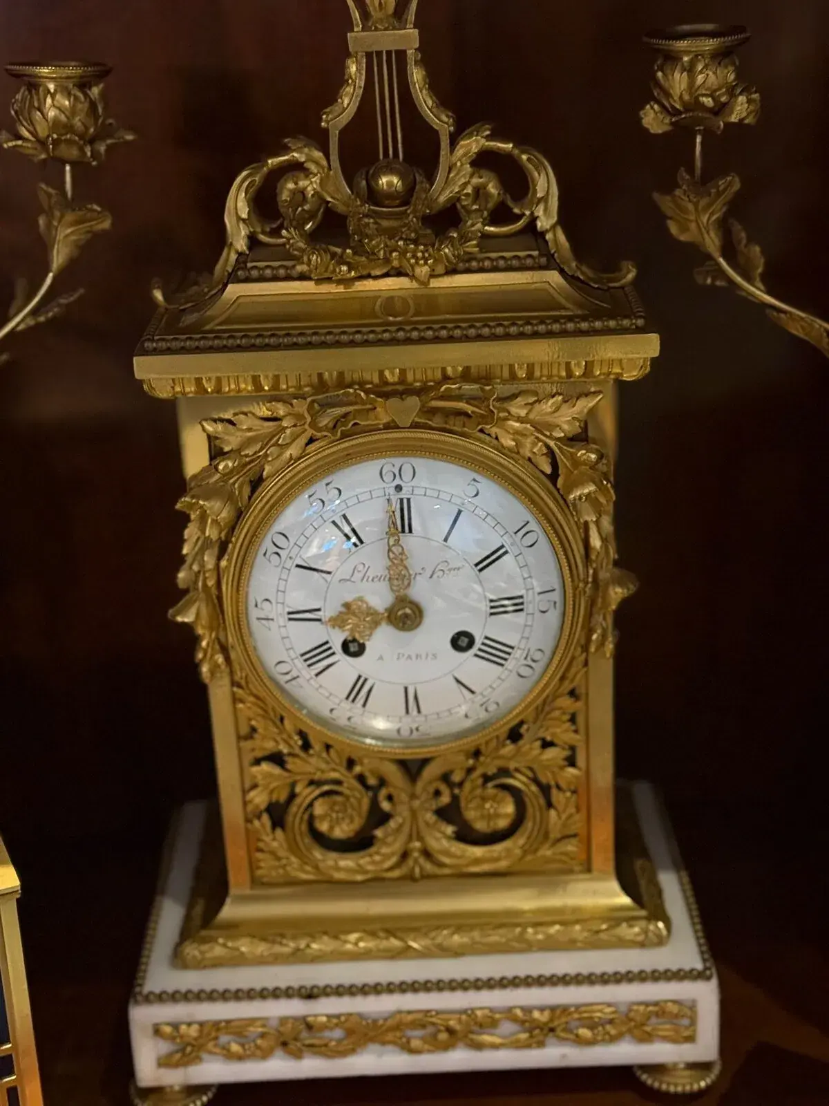Gros plan sur une horloge de cheminée Régulateur ornée de motifs floraux et de volutes, posée sur un socle en marbre blanc. Le cadran de l'horloge présente des chiffres romains, "Lhermitte Fils, Paris", et deux chandeliers flanquent cet objet d'atelier.
