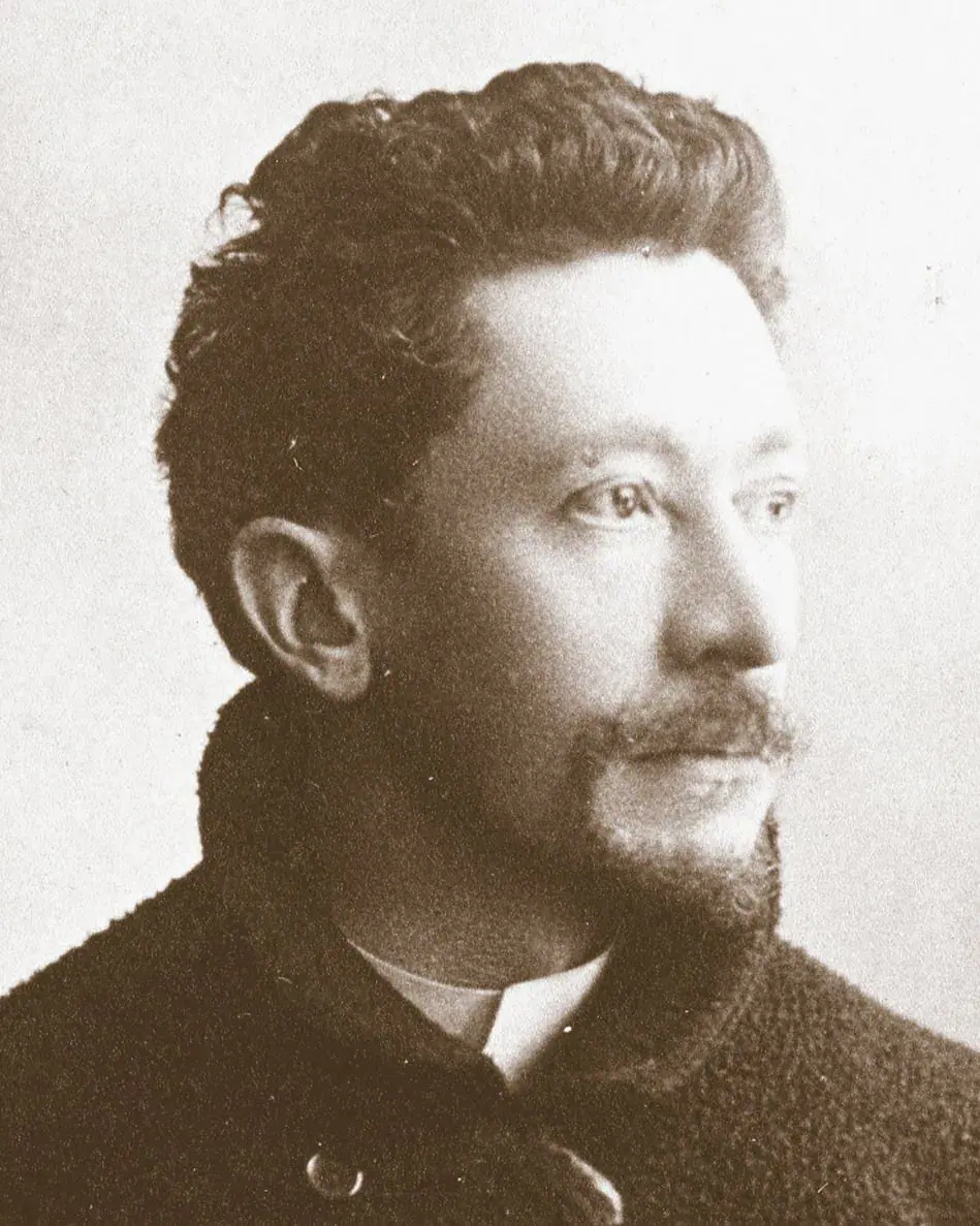 Portrait en tons sépia d'un homme aux cheveux noirs ondulés, portant une courte barbe et une moustache. Rappelant Emile Gallé, il porte un manteau sombre texturé et regarde légèrement vers la droite avec une expression calme et réfléchie sur un fond clair uni.