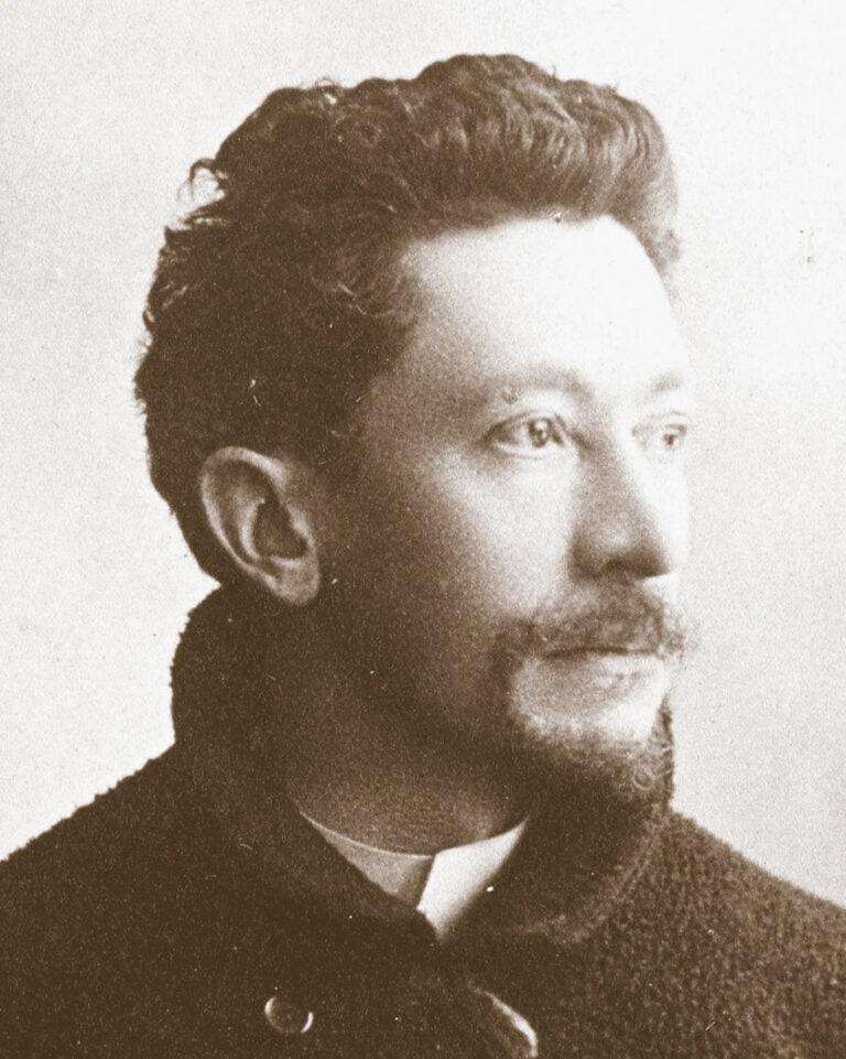 Portrait en tons sépia d'un homme aux cheveux noirs ondulés, portant une courte barbe et une moustache. Rappelant Emile Gallé, il porte un manteau sombre texturé et regarde légèrement vers la droite avec une expression calme et réfléchie sur un fond clair uni.