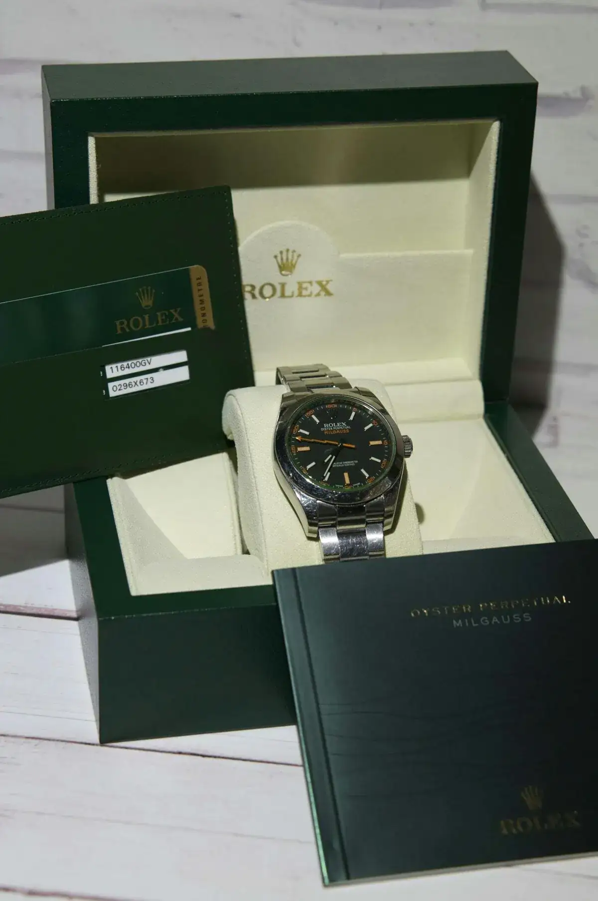 Une montre Rolex Oyster Perpetual Milgauss en acier inoxydable est présentée dans une boîte ouverte de couleur verte et crème. Des cartes Rolex vertes et un livret sont disposés autour de la boîte, présentant les détails d'authentification et de marquage de la montre. L'arrière-plan est une surface en bois blanc.