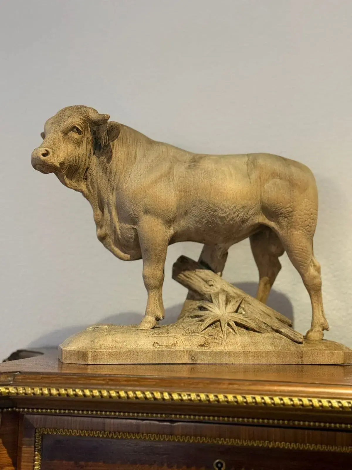 Une sculpture en bois détaillée d'un taureau, réalisée dans la tradition de Vallorbe, repose sur une base rectangulaire avec un tronc et une plante sculptés. Le taureau est tourné vers l'avant, avec des détails musculaires complexes, et repose sur une surface en bois poli avec des garnitures dorées.