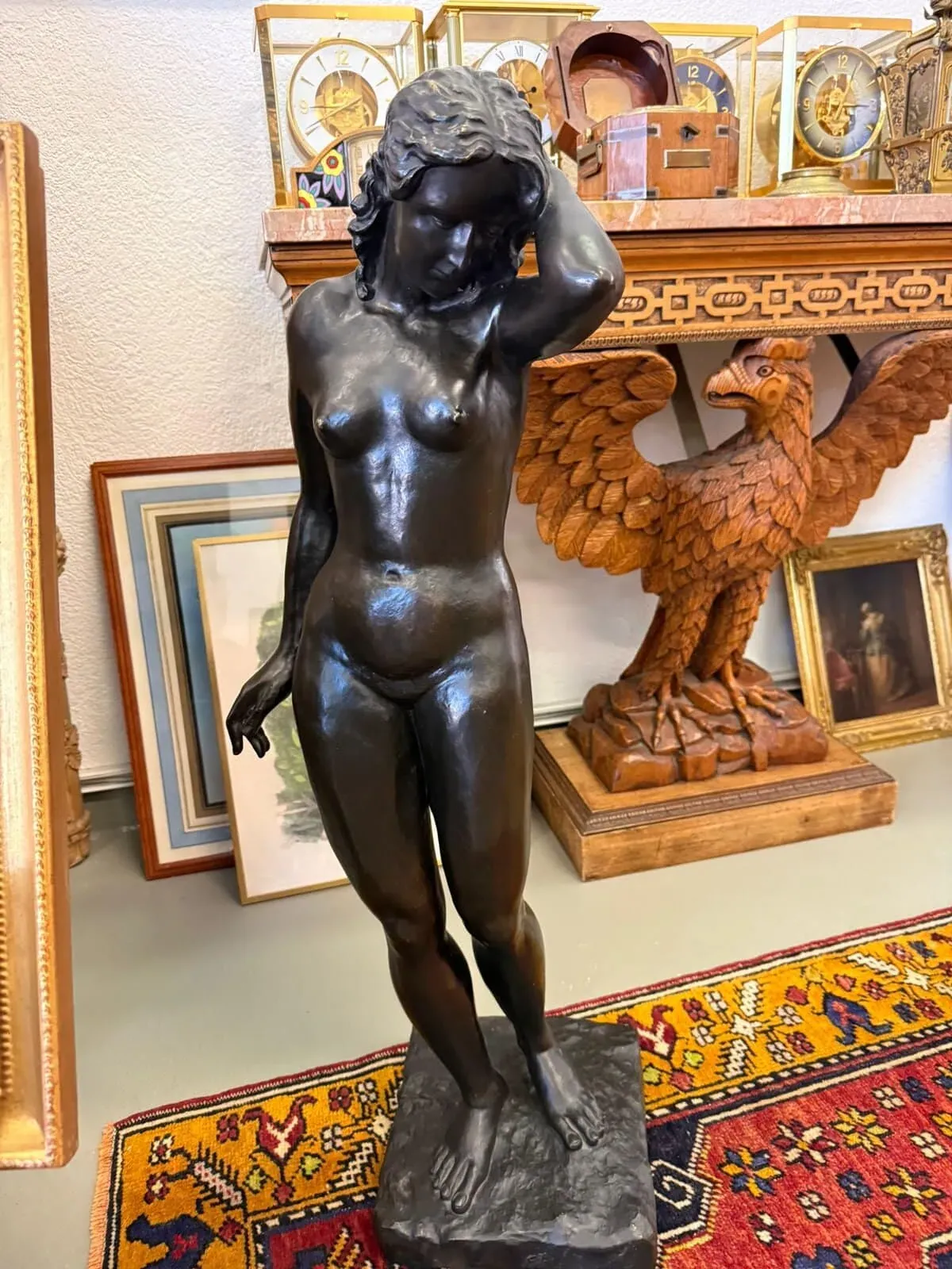 Une statue en bronze foncé d'une femme nue se dresse sur une base carrée à Thônex, le bras gauche replié derrière la tête. La statue est entourée d'œuvres d'art encadrées, d'horloges anciennes et d'un aigle en bois sculpté sur un tapis à motifs rouge et or.