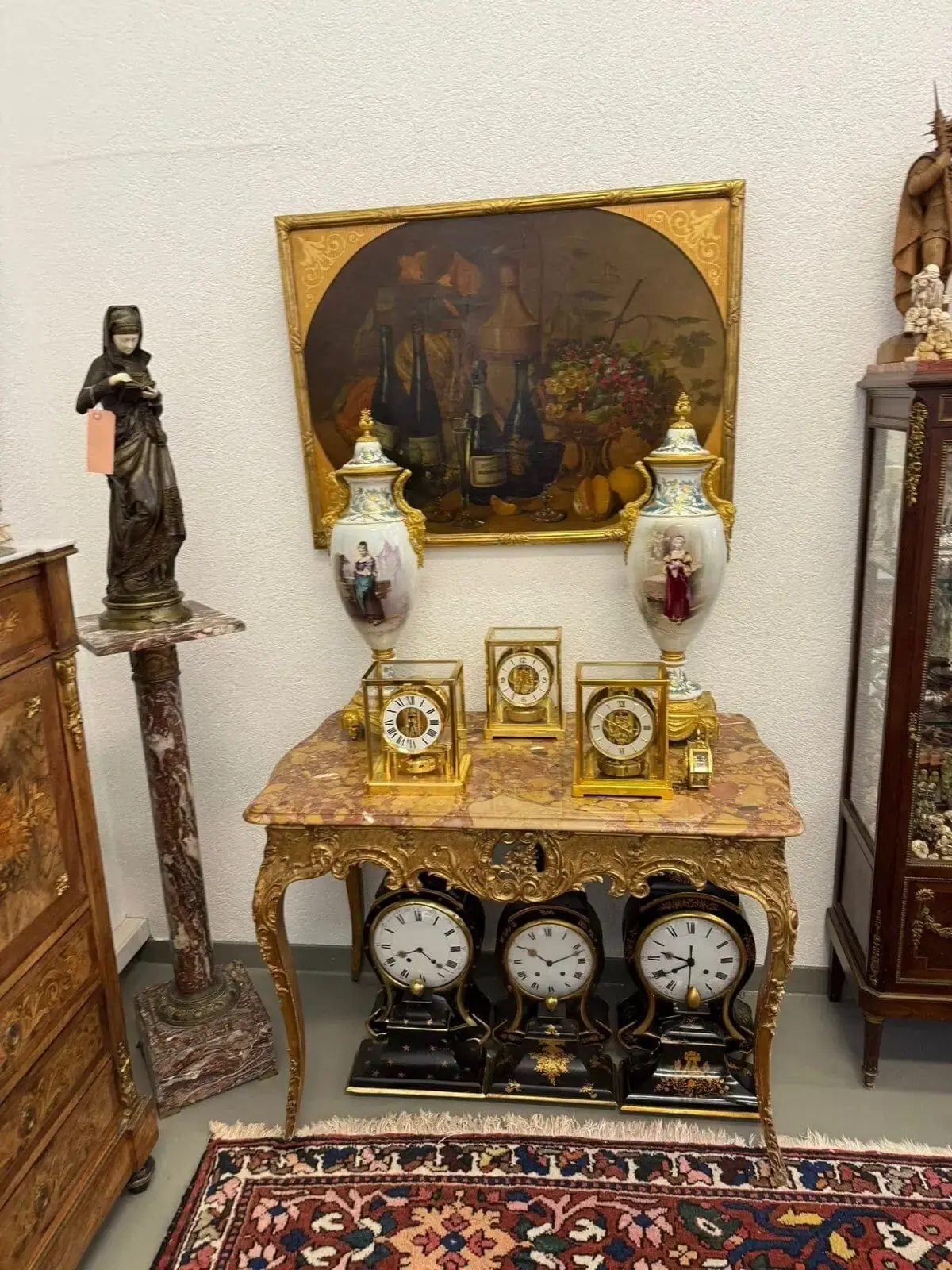Une pièce ancienne au décor orné : deux grands vases en porcelaine, cinq horloges en or et trois horloges noir et or sur une table en marbre. Derrière, une nature morte avec des fruits et des bouteilles ; à gauche, un piédestal en marbre avec une statue. Tapis oriental au sol.