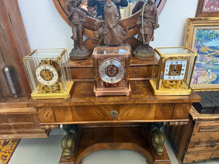 Trois horloges de cheminée ornées, dont une élégante Atmos Jaeger-LeCoultre, trônent sur une table en bois poli aux pieds incurvés. Derrière elles, un miroir rond, des statues en bronze et des œuvres d'art encadrées sur un mur blanc créent une exposition d'antiquités classique.