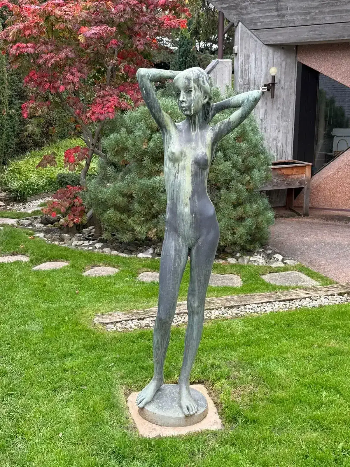 La statue en bronze d'une femme nue est posée sur l'herbe à Carouge, les mains derrière la tête. Derrière elle, des arbustes verts, un arbre à feuilles rouges, un chemin de pierre et un bâtiment avec un mur rose et une grande fenêtre : une scène de jardin accueillante.