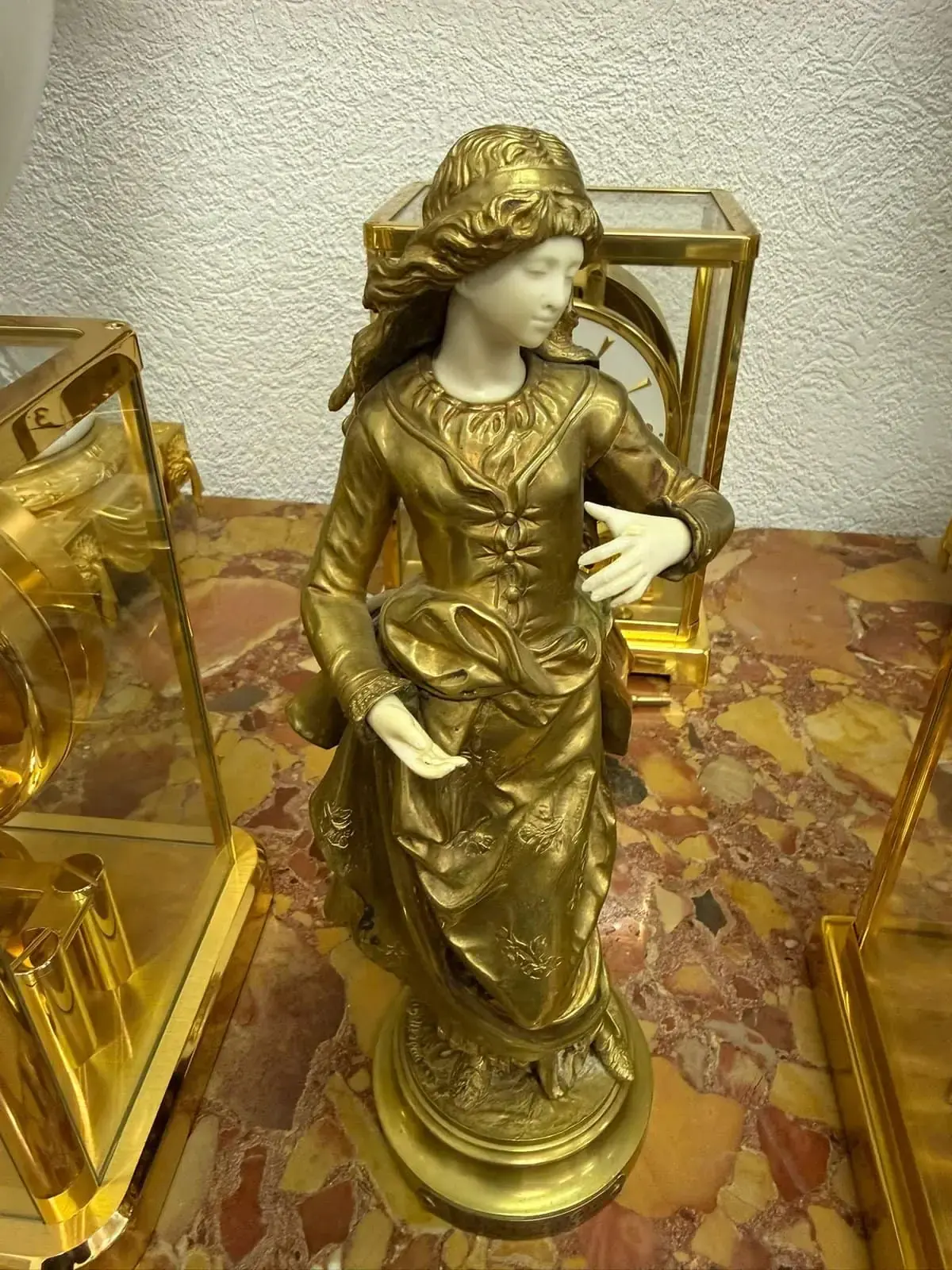 Une statue dorée et blanche d'une femme vêtue d'une robe fluide détaillée se dresse sur une surface de marbre marron et orange tacheté. Ses cheveux sont sculptés en vagues, son visage présente des détails délicats et des boîtiers d'horloge Bardonnex encadrés d'or sont visibles à l'arrière-plan.