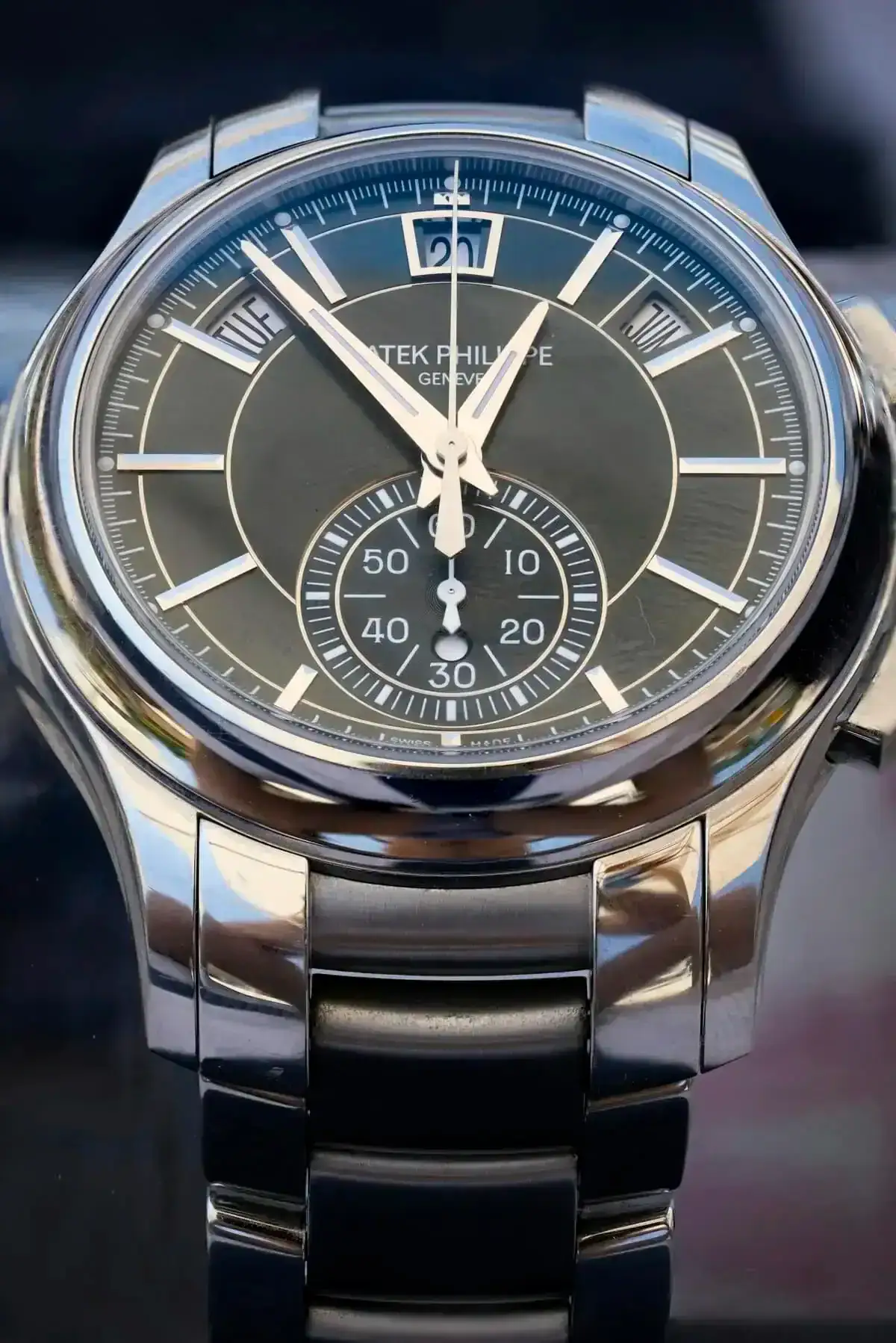 Gros plan sur une montre-bracelet de luxe Patek Philippe dotée d'un bracelet métallique. Le cadran noir de la montre présente des index argentés, un guichet de date à 12 heures et un cadran auxiliaire au-dessus de 6 heures indiquant les secondes. L'heure indique 11:58:00. La montre est très bien polie.