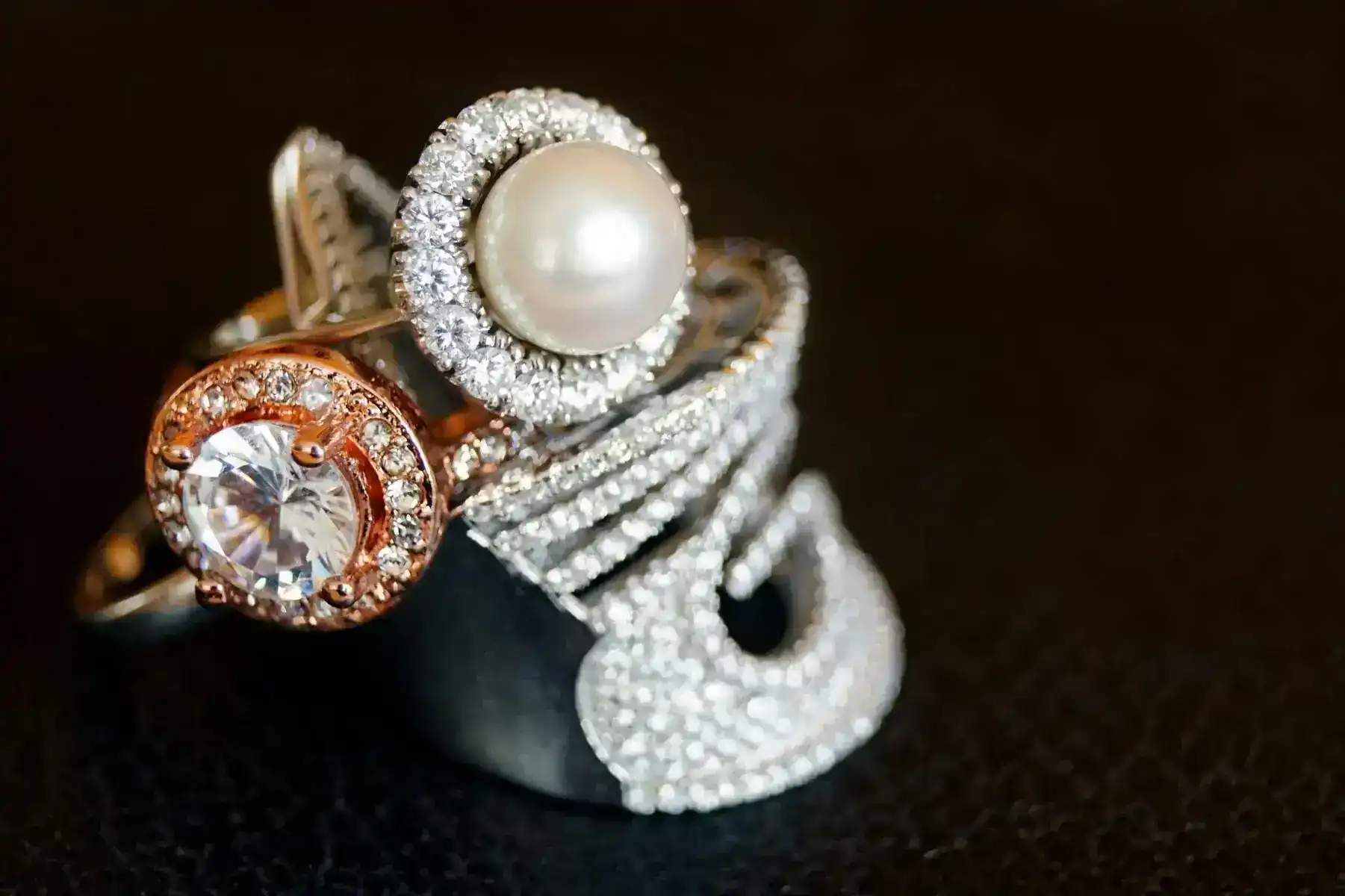Trois bagues ornées sont présentées sur une surface sombre et texturée. L'une présente une perle entourée de diamants, l'autre un diamant rond avec un halo en or rose, et la troisième est une large bande d'argent incrustée de petits diamants étincelants aux motifs complexes.