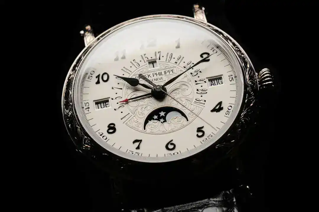 Gros plan sur une montre-bracelet de luxe Patek Philippe avec un cadran blanc, des chiffres arabes noirs, un affichage des phases de lune, des fenêtres pour le jour et le mois, et une gravure ornementale sur la lunette argentée. Le bracelet en cuir noir Patek Philippe est partiellement visible sur un fond sombre.